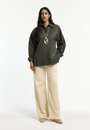 Frau trägt ein übergroßes, dunkelgrünes Hemd mit Knöpfen, beige Hosen mit weitem Bein, beige Schuhe und eine große, geometrische Anhängerkette, steht vor weißem Hintergrund.