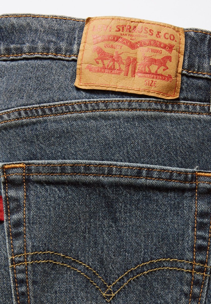 Jeans in denim blu con un'etichetta in pelle beige. L'etichetta presenta grafiche rosse di due cavalli, testo e dettagli di cucitura in arancione.