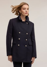 Abrigo de lana azul marino oscuro con un alto cuello, diseño de doble botonadura y botones dorados. Cuenta con bolsillos laterales y una tela texturizada.