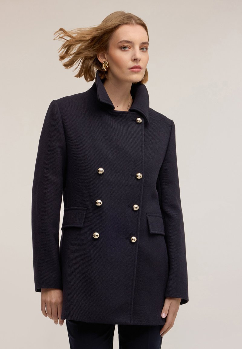 Abrigo de lana azul marino oscuro con un alto cuello, diseño de doble botonadura y botones dorados. Cuenta con bolsillos laterales y una tela texturizada.
