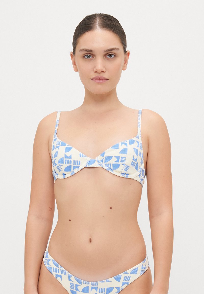 Krém alapon geometric kék mintával díszített bikini szett. A top állványos és állítható pántokkal rendelkezik; a bugyi alacsony derekú, ugyanazzal a mintázattal.