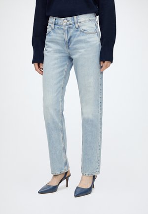 Femme portant un jean droit bleu clair, un haut à manches longues bleu marine et des talons slingback bleu marine à bout pointu, debout sur un fond uni.