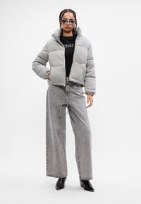 Kvinde iført grå pufferjakke, sort cropped top, brede grå jeans, sorte spidse støvler og solbriller med fletninger.