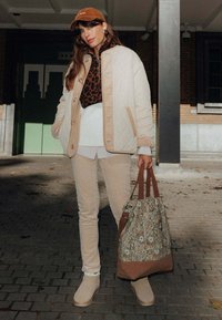Veste matelassée beige sur un pull imprimé léopard marron, associée à un pantalon clair, des bottes beiges et un sac fourre-tout fleuri. Casquette marron et tops superposés visibles.