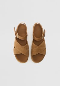 Sandalias de ante marrón con tiras cruzadas y cierres de hebilla ajustables, suelas planas y el logo de UGG grabado en las plantillas.