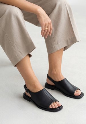 Person iført beige cropped bukser og sorte læder sandaler med åben tå og ankelrem, siddende med den ene hånd hvilende nær knæet.