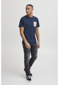 T-shirt in cotone blu navy con taschino a fantasia multicolore, abbinato a jeans grigi e sneaker nere. Design semplice e casual.