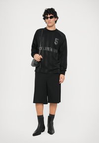 Svart langermet genser med grafisk trykk og nummer 5, paret med svarte shorts og elegante svarte ankelstøvler, holder en liten veske.