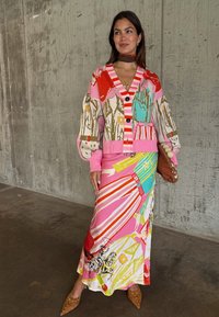 Γυναίκα που φοράει πολύχρωμη ζακέτα με σχέδια και ταίριαστο μακρύ φούστα, κρατώντας ένα καφέ τσαντάκι χειρός, στέκεται μπροστά σε φόντο από τσιμεντένιο τοίχο.