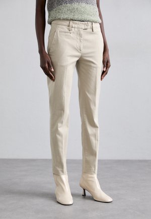 Osoba ubrana w beżowe, dopasowane spodnie w zestawieniu z kremowymi botkami na obcasie, stojąca na szarej podłodze na tle jasnoszarej ściany.