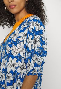 Vestido floral azul com flores brancas e pretas, decote laranja, mangas curtas bufantes e um tecido texturizado com detalhes de franzido.