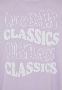 Světle levandulové tričko se velkým bílým textem ve vlnovém písmu, s nápisem "URBAN CLASSICS" opakovaným dvakrát ve dvou řadách.