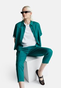 Zestaw z niebiesko-zielonej lnianej koszuli i spodni, z białą koszulką pod spodem. Uzupełniony czarnymi okularami przeciwsłonecznymi i plecionymi czarnymi butami.