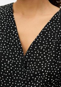 Mustarakk mustriga must blouse, valgete ebakorrapäraste täppidega, V-kaelus ja nööbid rinnal. Pehme tekstuur ja juhuslik disain.