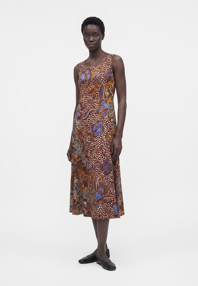 Γυναίκα που φοράει καφέ midi φόρεμα χωρίς μανίκια με μπλε και πορτοκαλί floral και πουά σχέδια, σε συνδυασμό με μαύρα επίπεδα παπούτσια, που στέκεται σε λευκό φόντο.