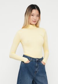 Top amarillo claro de manga larga con cuello alto, combinado con unos vaqueros de mezclilla azul de tiro alto. La blusa tiene una textura suave y un diseño ajustado.