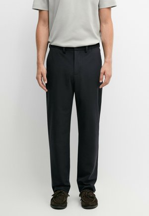 Uomo che indossa pantaloni neri a gamba dritta, una camicia grigia chiara a maniche corte e scarpe stringate marrone scuro, in piedi davanti a uno sfondo semplice.