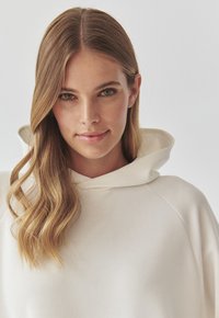 Felpa beige chiaro con una texture morbida, un grande cappuccio e maniche raglan. Il collo è rotondo e liscio, fondendosi con il tessuto.
