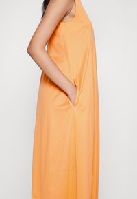Robe orange sans manches avec poche latérale, la main droite du mannequin placée à l'intérieur de la poche, visible des épaules jusqu'à mi-cuisse.
