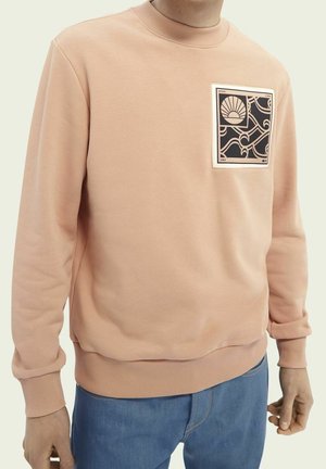 Mann trägt ein helles pfirsichfarbenes Sweatshirt mit einem schwarzen und beige abstrakten quadratischen Motiv auf der oberen linken Brust, kombiniert mit blauen Jeans.