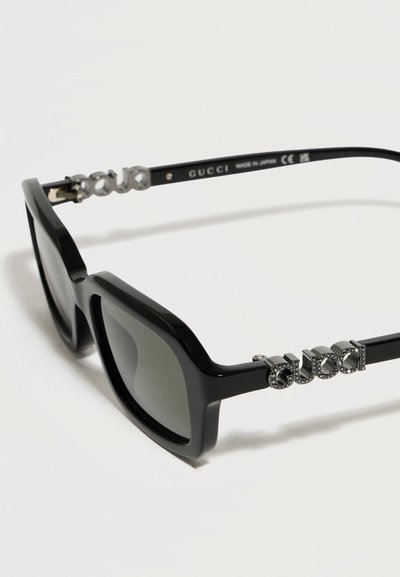 Lunettes de soleil rectangulaires noires Gucci avec verres gris et détail logo argenté orné de petits strass sur les branches.