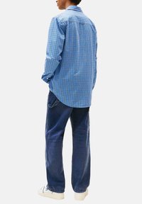 Uomo in piedi di spalle, che indossa una camicia a quadri blu a maniche lunghe, jeans blu scuro e sneaker bianche su uno sfondo neutro.