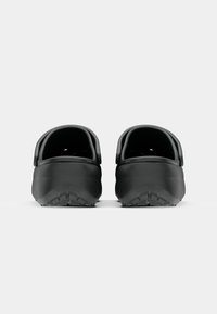 Paire de sabots noirs sans lacets avec semelles texturées, montrée de dos sur un fond gris clair uni.