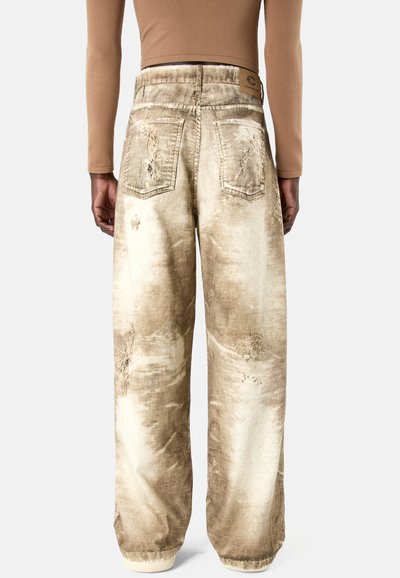 Personne portant un pantalon en velours côtelé beige usé, coupe large, avec poches arrière et texture délavée, vue de dos.