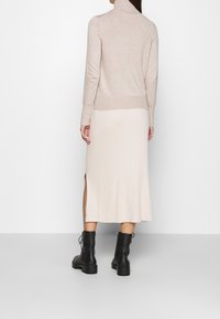 Femme portant un pull à col roulé beige, une jupe midi côtelée crème avec une fente, et des bottines noires à lacets, debout devant un fond blanc.