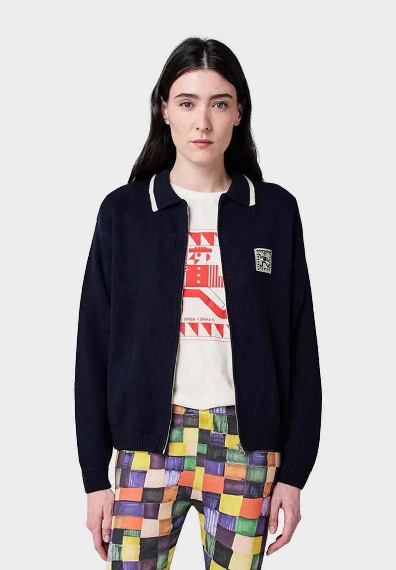 Kvinna som bär en marinblå zip-up-tröja över en vit grafisk T-shirt och färgglada patchwork-mönstrade byxor, stående mot en ensfärgad bakgrund.