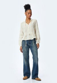 Camicia crema con bottoni, vita peplum, tessuto testurizzato e maniche lunghe, abbinata a jeans in denim a gamba larga.
