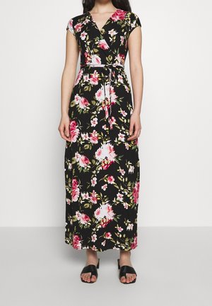 Vrouw draagt een zwarte bloemenprint wikkel maxi-jurk met korte mouwen en een gestrikte taille, gecombineerd met zwarte open-toe sandalen met hak.