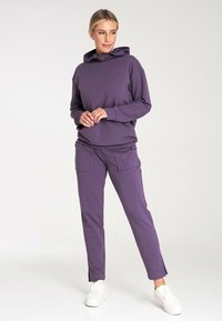 Fioletowa bluza z kapturem na sznurki, połączona z dopasowanymi fioletowymi spodniami ze zwężanymi nogawkami i bocznymi kieszeniami, noszonymi z białymi sneakersami.