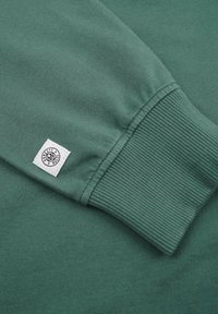 Grön bomulls sweatshirt med ribbad muddDetalj och en kvadratisk etikett med en logotyp. Slät textur, enkel design.