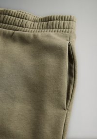 Khaki värvi dressipüksid, millel on tekstureeritud ja pehme kude; elastse vöökoha ja küljetaskutega. Suurendatud vaade, mis rõhutab materjali ja väljanägemist.