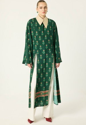 Femme portant une longue tunique verte à motifs avec un large col beige, un pantalon large blanc et des chaussures rouges pointues, debout devant un fond uni.