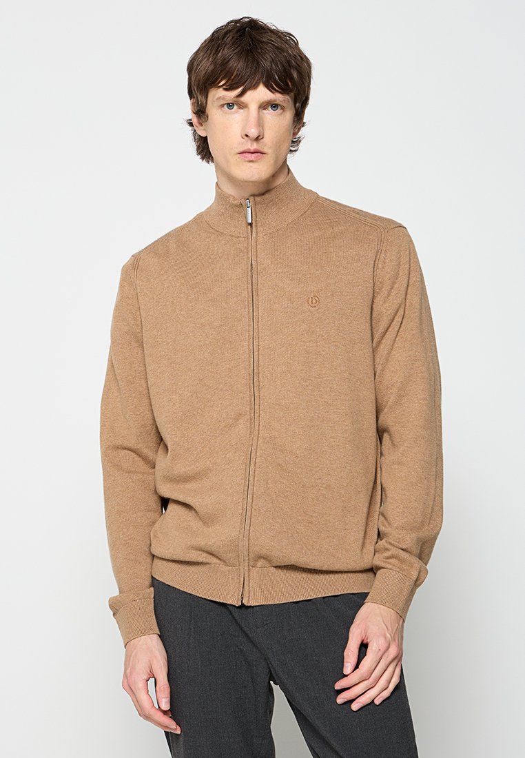 Mann in einem hellbraunen Reißverschluss-Pullover und dunkelgrauen Hosen, der vor einem schlichten hellen Hintergrund steht.