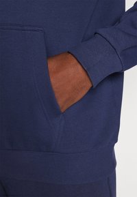 Mørkeblå sweatshirt med kængurulomme, ribbede manchetter og ribkant. Glat tekstur med flade sømme og kontrasterende syning.