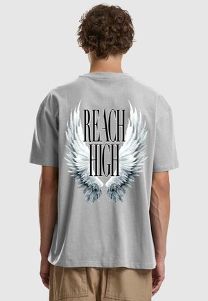 Sivé bavlnené tričko s veľkým bielym motívom krídel a čiernym textom "REACH HIGH" na chrbte. Krátke rukávy a voľný strih.