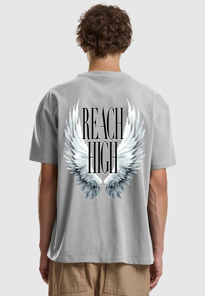 Γκρι βαμβακερό T-shirt με μεγάλο λευκό σχέδιο φτερών και μαύρο κείμενο που αναγράφει "REACH HIGH" στην πλάτη. Κοντά μανίκια και άνετη εφαρμογή.