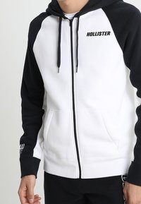 Herrarnas zip-up hoodie i vitt och svart. Tillverkad av mjukt material, har kontrasterande ärmar, framfickor och "HOLLISTER"-logotyp på bröstet.