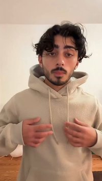 Lysebeige fleece-hættetrøje med forlomme, snoretræk og blød tekstur. Personen har mørkt, bølget hår og befinder sig indendørs.