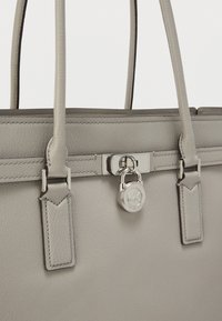 Bolso de cuero gris texturizado con herrajes plateados, que presenta un detalle de candado y dos asas robustas. Diseño minimalista con acentos cosidos.