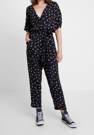 Vrouw draagt een zwarte jumpsuit met witte bladprints en een tailleband, gecombineerd met marineblauwe hoge sneakers.