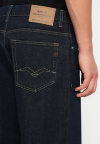 Mørkeblå denimjeans med en teksturert finish. Har et synlig lærmerke og et unikt hvitt søm mønster på baklommen.