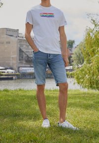 Camiseta blanca con el logotipo multicolor de "Pepe Jeans", combinada con pantalones cortos de mezclilla azul claro y zapatillas blancas, ubicada en un área exterior con césped.