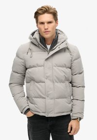 Chaqueta acolchada gris claro con capucha, que presenta un frente con cremallera, puños elásticos y un diseño acolchado. El material parece suave e insulated.