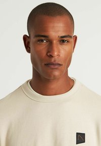 Sudadera de algodón beige con cuello redondo, que presenta un parche negro con un diseño angular en naranja en el pecho. Textura suave, estilo minimalista.