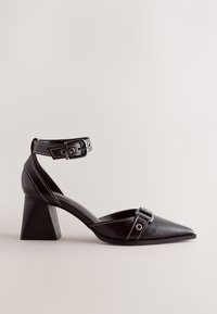 Chaussure à talon en cuir noir pour femme, à bout pointu, bride cheville avec boucle et talon bloc sur fond neutre.