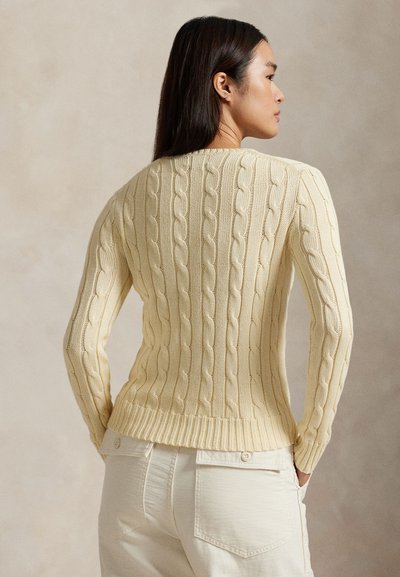 Donna con capelli lisci e scuri che indossa un maglione beige chiaro a trecce e pantaloni bianchi, in piedi con le mani nelle tasche, vista di profilo.
